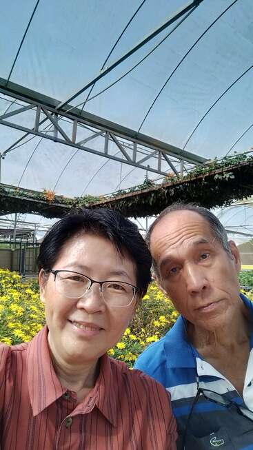 Das Bild zeigt ein Selfie von einem asiatischen Paar in einem Gewächshaus mit gelben Blumen und einer Metallkonstruktion im Hintergrund.