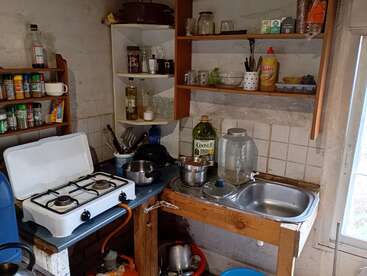 Cette image montre une petite cuisine rustique avec une cuisinière à gaz, des étagères ouvertes, divers ustensiles, des pots, des bocaux, des épices, un évier et un désordre général.