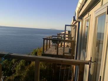 The image depicts a house with a balcony overlooking the ocean, featuring a wooden deck and a white downspout. The scene is set against a clear blue sky.