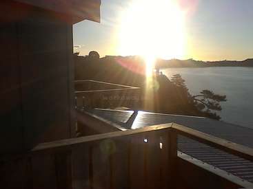 The image depicts a serene scene of a house\'s deck overlooking a body of water, with the sun shining brightly in the background.