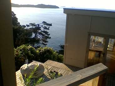The image depicts a serene scene of a house with a deck overlooking a body of water, surrounded by lush greenery and trees.
