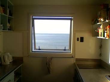 The image depicts a kitchen with a window overlooking the ocean, featuring a kettle, sink, and shelves stocked with various items.