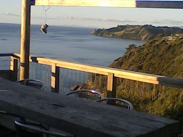 The image depicts a wooden deck with a railing overlooking a body of water and a rocky coastline, featuring a table and chairs in the foreground.