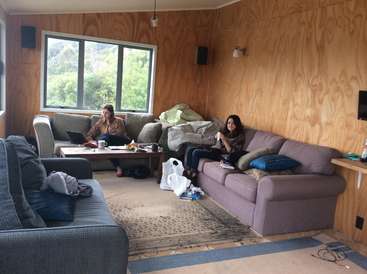 The image depicts a cozy living room with two women seated on couches, surrounded by a warm and inviting atmosphere. The room features wooden walls and a large window.