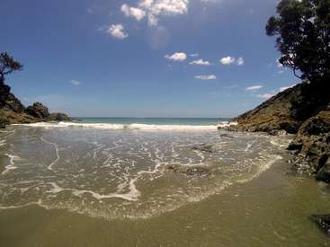 The image depicts a serene beach scene with a sandy shore, gentle waves, and rocky outcroppings, set against a clear blue sky with scattered clouds.