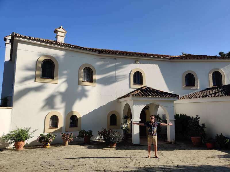 Ein Mann steht mit ausgestreckten Armen vor einer weißen Villa mit Bogenfenstern, Topfpflanzen und einem strahlend blauen Himmel über ihm.