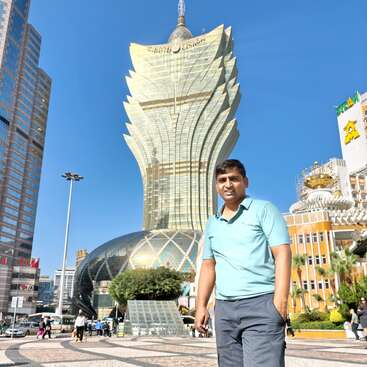 Ein Typ in einem hellblauen Hemd steht auf einem gemusterten Platz, und das berühmte Grand Lisboa Hotel in Macau ragt beeindruckend in den strahlend blauen Himmel.