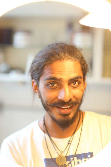 Un homme souriant aux cheveux longs et à la moustache frisée porte des colliers et une chemise de couleur claire, avec un arrière-plan intérieur flou et un éclairage doux.