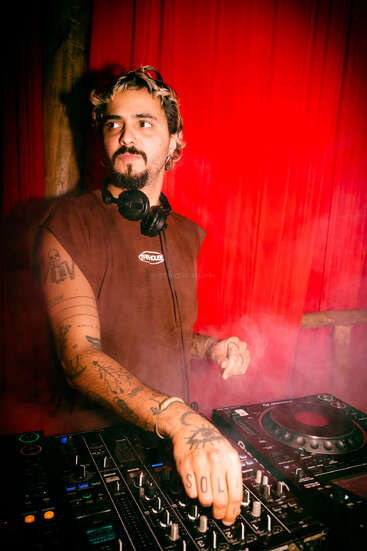 Ein DJ steht an einem Mischpult, umgeben von roten Vorhängen und Rauch. Er hat Tattoos, trägt Kopfhörer um den Hals und schaut nachdenklich zur Seite.