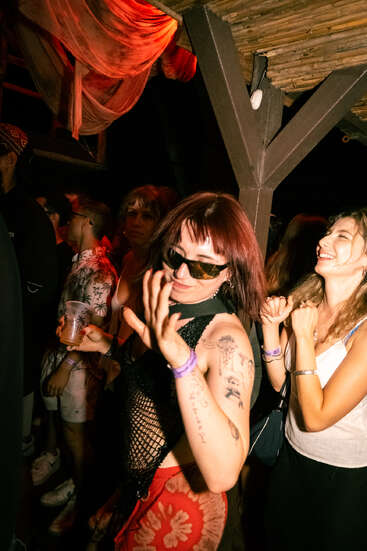Eine Frau mit Tattoos und Sonnenbrille tanzt auf einer ausgelassenen Party in einem Innenraum, ein Getränk in der Hand, umgeben von lächelnden Freunden unter warmer, bunter Beleuchtung und rustikalem Dekor.