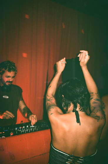 Eine Frau mit Tattoos tanzt mit erhobenen Armen vor einem DJ, der Musik auflegt. Der Hintergrund ist schwach beleuchtet und mit rot-orangefarbenen Vorhängen versehen, was eine lebhafte Atmosphäre schafft.