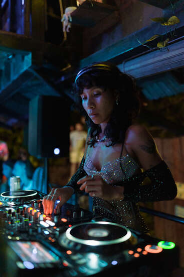 Eine DJane in glitzerndem Outfit legt an einem DJ-Controller in einem schummrig beleuchteten Lokal Musik auf. Bunte Lichter sorgen um sie herum für eine lebhafte und energiegeladene Atmosphäre.