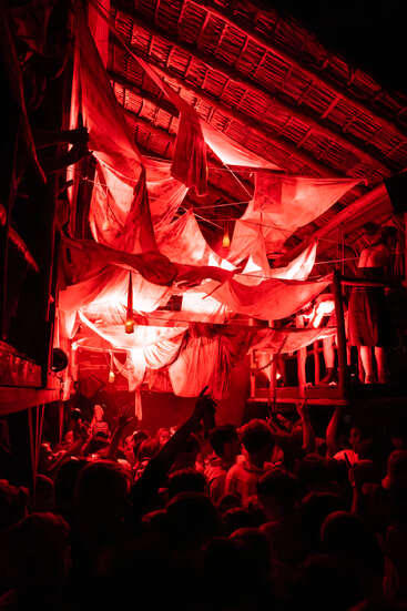 Eine überfüllte Partyszene in einem Innenraum, beleuchtet von dramatischem rotem Licht. Stoffvorhänge hängen von der Decke, während die Leute darunter energiegeladen tanzen und eine intensive, pulsierende Atmosphäre schaffen.