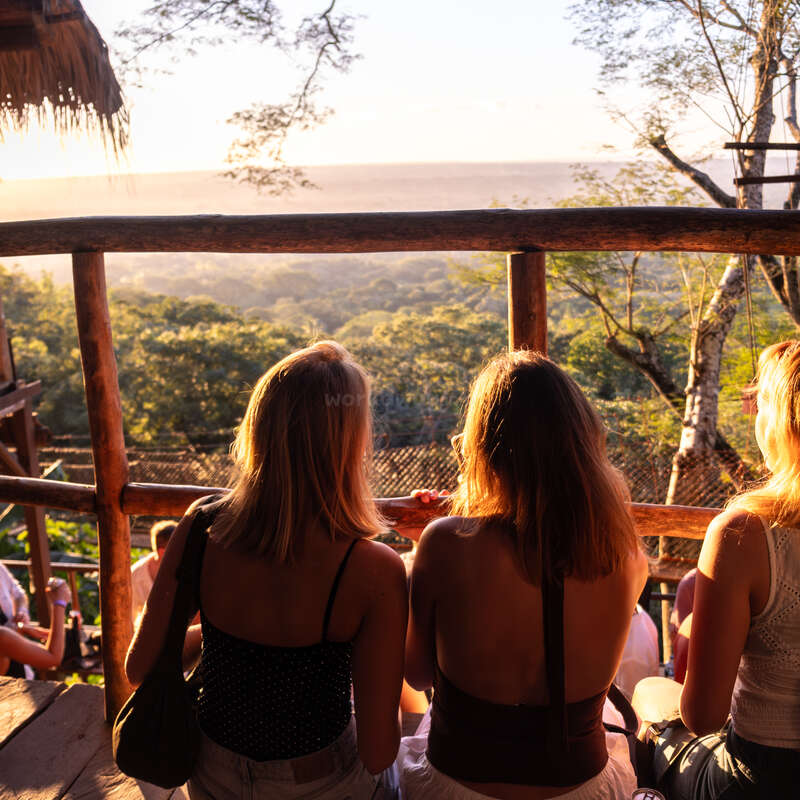 Drei Frauen sitzen auf einem Holzbalkon und blicken auf einen üppig grünen Wald und ferne Hügel, während der goldene Sonnenuntergang alles in warmes, ruhiges Licht taucht.