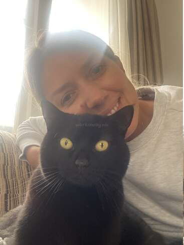 L'image montre une femme souriante avec un chat noir devant elle, assise sur un canapé devant une fenêtre avec des rideaux.