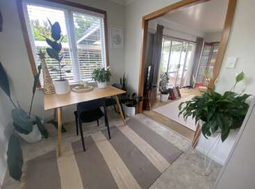 L'image représente une pièce confortable avec un bureau, des plantes et un tapis, avec une porte donnant sur une autre pièce avec une guitare et d'autres plantes, dans une maison.