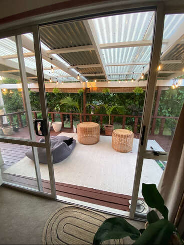 L'image représente un espace intérieur-extérieur serein, avec une porte coulissante en verre qui s'ouvre sur une terrasse avec un tapis blanc, des meubles en osier et de la verdure luxuriante, créant ainsi une ambiance paisible.