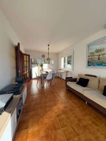 Ein helles Wohn-Esszimmer mit braunem Fliesenboden, modernen Möbeln, einem beigen Sofa, einem weißen Esstisch, großen Fenstern, Wandbildern und dekorativen Akzenten überall.