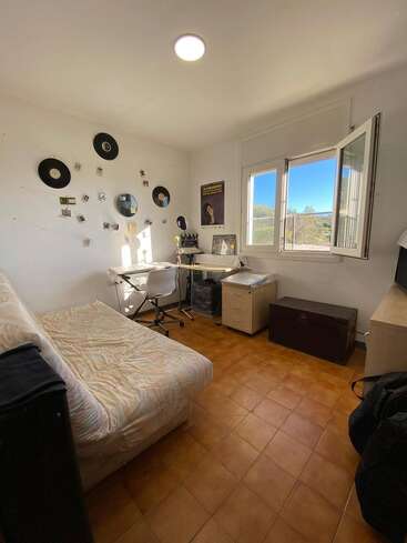 Das ist ein aufgeräumtes, sonnendurchflutetes Schlafzimmer mit einem Bett, einem Schreibtisch, einem Bürostuhl, an der Wand hängenden Schallplatten, Postern, einem offenen Fenster und einem warmen Terrakotta-Fliesenboden.