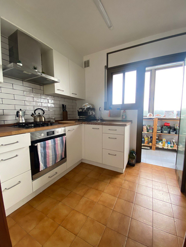 Dieses Bild zeigt eine helle Küche mit weißen Schränken, Arbeitsplatten aus Holz, einem Herd, einem Backofen, einer gefliesten Küchenrückwand, einem großen Fenster, einem hellbraunen Fliesenboden und einem angrenzenden Vorratsbereich.