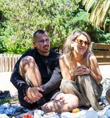 Ein Mann und eine Frau sitzen draußen auf einer Decke und genießen ein sonniges Picknick mit Essen und Getränken. Sie wirken entspannt, lächeln und sind von üppigem Grün umgeben.