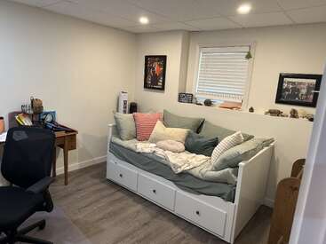 This cozy room features a daybed with pastel pillows, a wooden desk with supplies, an office chair, wall art, decorations, and a window with blinds.