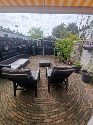 La imagen muestra un acogedor patio con dos sillas, un sofá y una mesa, rodeado de macetas y un pequeño cobertizo al fondo, bajo un toldo a rayas.