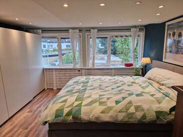 La imagen muestra un dormitorio con una cama grande, paredes blancas y una ventana que da a una calle, con un edredón de motivos geométricos y un cuadro en la pared.
