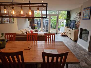 La imagen muestra un salón moderno con mesa de comedor, sofá, televisor y chimenea, con un gran ventanal y una puerta corredera de cristal que da a un patio trasero.