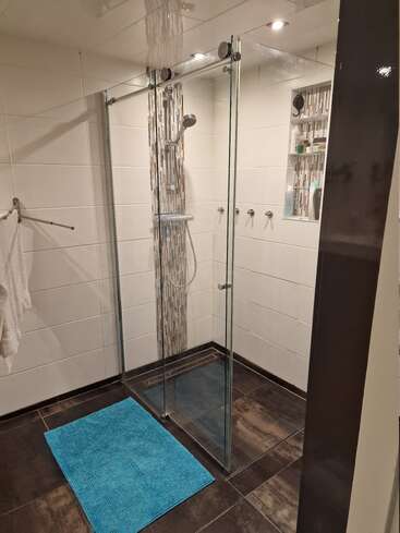 La imagen muestra un cuarto de baño moderno con mampara de ducha de cristal, con una alfombrilla azul en el suelo, baldosas blancas y suelo de madera oscura, que desprende un ambiente limpio y minimalista.