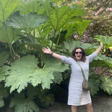 Une femme joyeuse vêtue d'une robe légère se tient les bras tendus, souriante devant d'énormes feuilles vertes, entourée de plantes et d'arbres luxuriants.