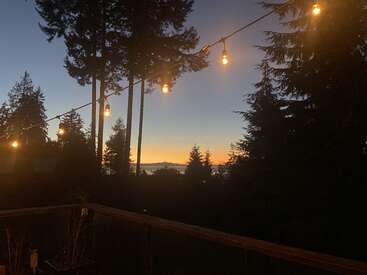 À la tombée de la nuit, des guirlandes lumineuses brillent chaleureusement au-dessus d'une terrasse en bois, entourée de grands arbres. Le coucher de soleil crée un ciel tranquille et coloré au-dessus de l'eau lointaine.