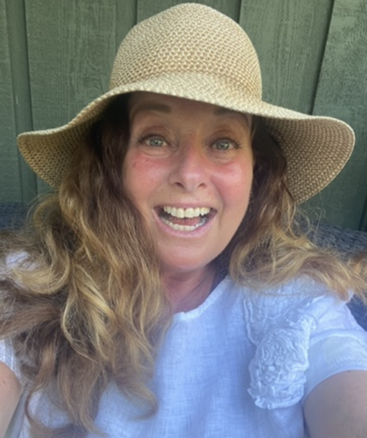 Une femme souriante aux longs cheveux ondulés porte un chapeau de paille à large bord et un haut blanc à motifs floraux, posant devant une clôture verte.