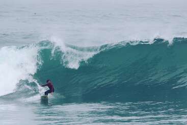 A person is surfing on a wave, with the ocean visible in the background. The water is clear and blue, and the sky is not visible. The overall atmosphere is one of excitement and adventure.