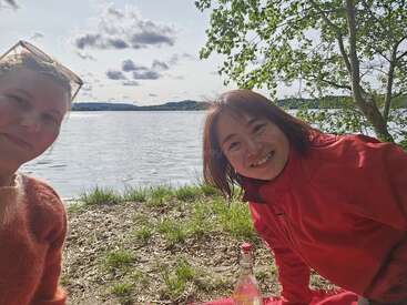 Zwei Frauen lächeln am Seeufer und sitzen auf dem Rasen in der Nähe eines Baumes. Auf einer roten Picknickdecke steht eine Flasche, hinter ihnen sieht man ruhiges Wasser und Wolken.