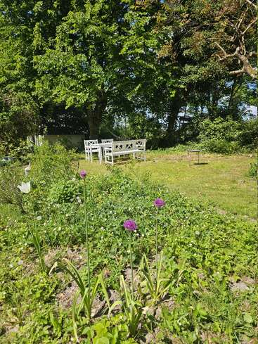 Eine wunderschöne Gartenszene mit grünen Bäumen, bunten Blumen und saftigem Gras. Weiße Gartenmöbel stehen im Schatten und laden zum Entspannen oder zu geselligen Runden ein.