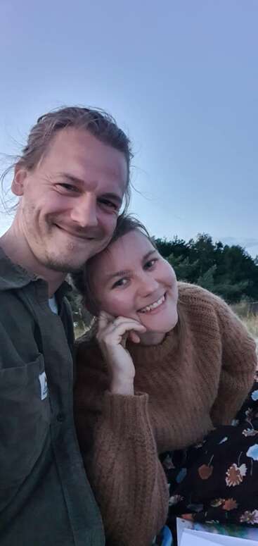 Ein lächelndes Paar macht draußen ein Selfie aus nächster Nähe, mit viel Grün im Hintergrund und einem strahlend blauen Himmel über ihnen; die beiden wirken glücklich und entspannt in ihrer Freizeitkleidung.