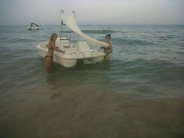 The image depicts two women in bikinis standing in the water next to a white boat with a slide, set against a gray sky.