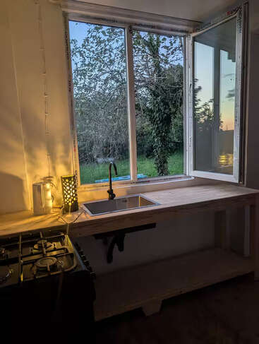 Eine gemütliche Küche mit Holzarbeitsplatte, Spüle und Herd. Durch das große, offene Fenster blickt man bei Sonnenuntergang auf einen üppig grünen Garten. Sanfte Stimmungsbeleuchtung sorgt für eine warme Atmosphäre.