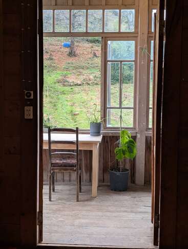Ein Holzraum mit einem schlichten Stuhl und Tisch vor großen Fenstern, Topfpflanzen, Blick auf einen grünen Hang, friedliche Atmosphäre, rustikales und gemütliches Interieur.