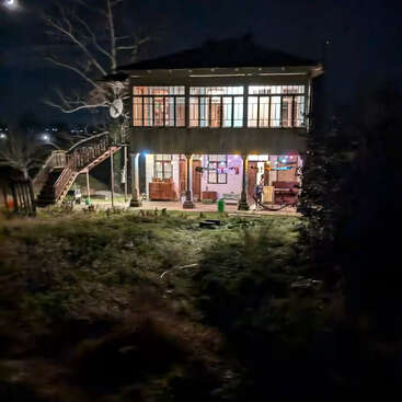 Uma casa de dois andares, bem iluminada à noite, tem janelas amplas, uma escada externa, luzes coloridas e um jardim gramado, criando uma atmosfera acolhedora e convidativa na escuridão.