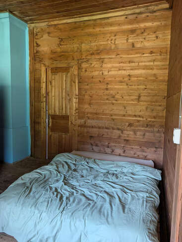 Dieses Bild zeigt ein gemütliches Holzzimmer mit einem ungemachten Bett und hellblauer Bettwäsche. Es gibt eine geschlossene Holztür und einen türkisfarben gestrichenen Wandabschnitt.