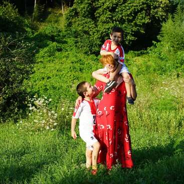 Eine Frau in einem roten Blumenkleid steht auf einer sonnigen Wiese und lächelt zwei Jungen in weißen Fußballtrikots an, von denen einer auf ihren Schultern sitzt.