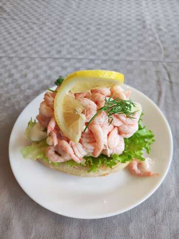 L'image représente un sandwich aux crevettes dans une assiette blanche, avec de la laitue, du citron et de l'aneth, sur une nappe grise au subtil motif à carreaux.