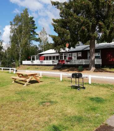 L'image représente une scène sereine d'une maison rouge avec un porche blanc, entourée d'arbres et d'une zone herbeuse avec une table de pique-nique et un grill.
