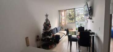 The image depicts a living room with a Christmas tree, a couch, and a dining table, featuring white walls and a window with a view of trees outside.