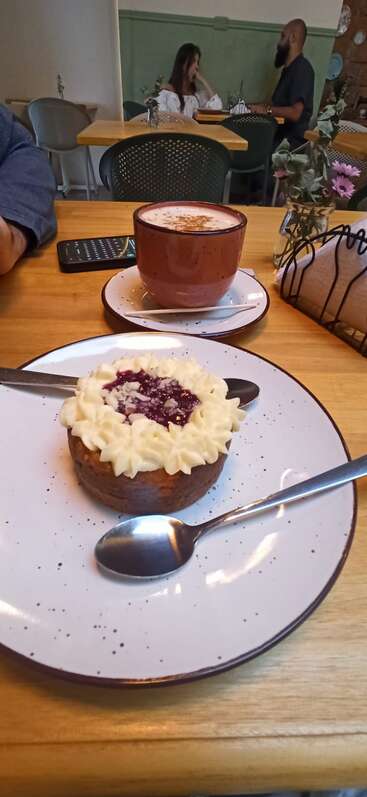 The image depicts a wooden table with a plate of dessert and a cup of coffee, set against a background of a restaurant with patrons seated at tables.