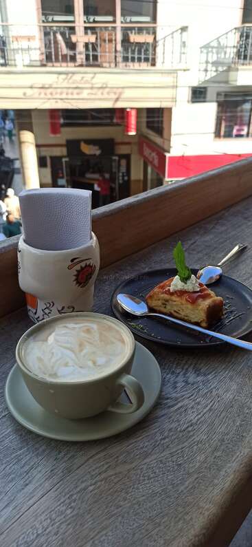 The image depicts a serene outdoor setting, featuring a cup of coffee and a slice of cake on a wooden table, with a building in the background.