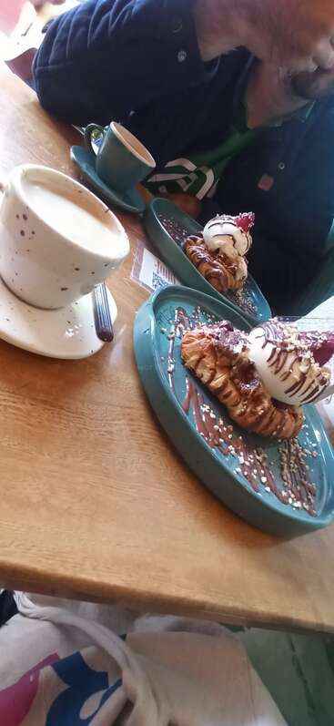 The image depicts a man sitting at a table with two plates of dessert and two cups of coffee, with a person's leg visible in the foreground.