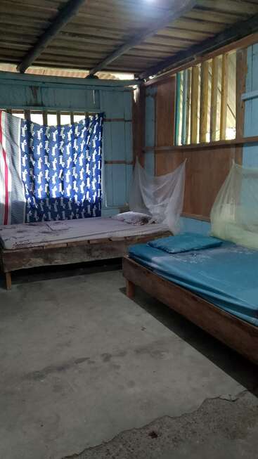Cette image montre une chambre simple avec deux lits, des moustiquaires, un sol en béton, des murs en bois bleu et un rideau bleu couvrant la fenêtre pour plus d'intimité.